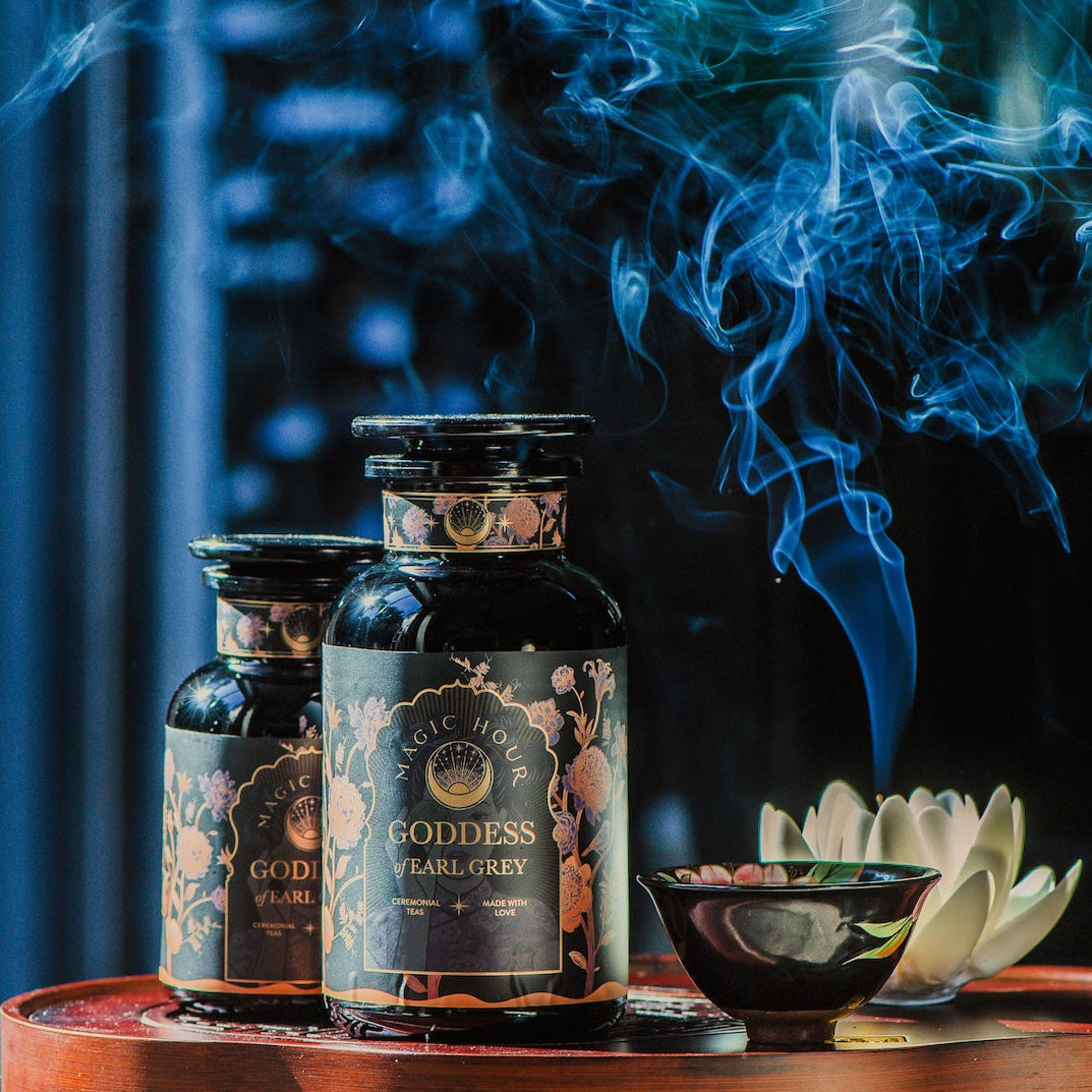 Two dark glass jars labeled "Goddess of Earl: Lady Luck Oriental Beauty Oolong Tea" from Magic Hour are placed on a wooden surface. A small bowl releases smoke, contributing to the serene ambiance. The background is softly blurred, accentuating the aromatic setting of this organic loose leaf tea captured in the image.