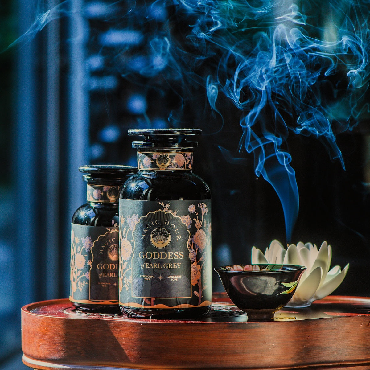 Two dark glass jars labeled &quot;Goddess of Earl: Lady Luck Oriental Beauty Oolong Tea&quot; from Magic Hour are placed on a wooden surface. A small bowl releases smoke, contributing to the serene ambiance. The background is softly blurred, accentuating the aromatic setting of this organic loose leaf tea captured in the image.