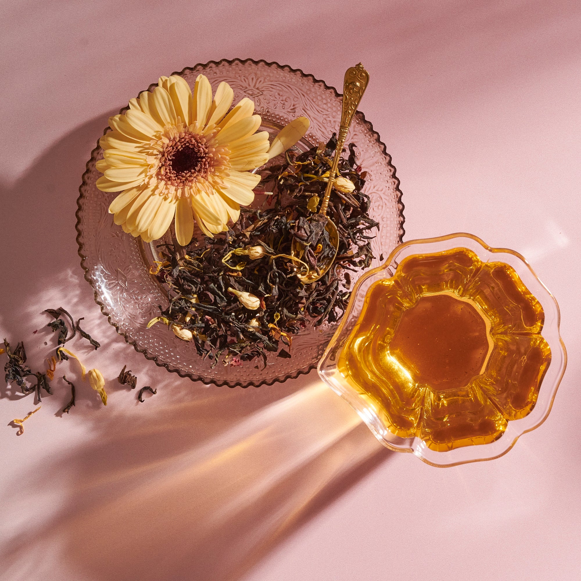 The Tea Discovery Club glass plate features loose curated tea blends, a yellow flower, and a gold spoon, set next to a cup of amber tea on a pink surface with sunlit shadows.