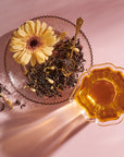 A glass dish filled with dried Goddess of Earl: Lady Luck Oriental Beauty Oolong Tea leaves by Magic Hour and a yellow flower sits next to a cup of brewed tea. A gold spoon rests on the tea leaves, releasing hints of bergamot. The scene is set against a pink surface, with light casting soft shadows.