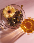 The Tea Discovery Club glass plate features loose curated tea blends, a yellow flower, and a gold spoon, set next to a cup of amber tea on a pink surface with sunlit shadows.