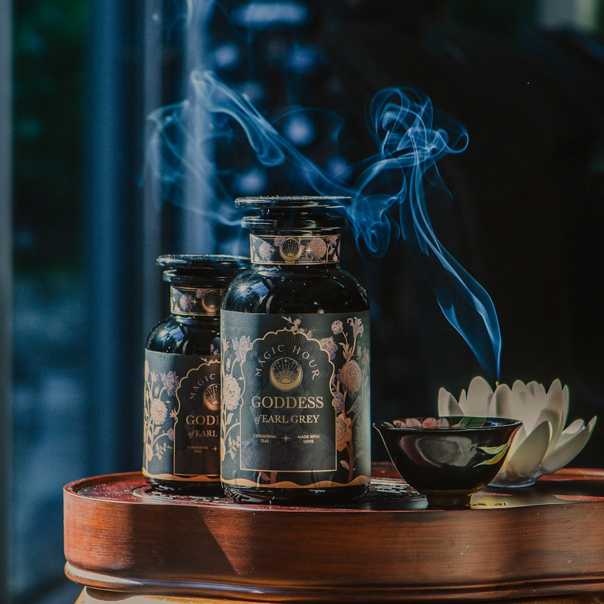 Two dark glass bottles of &quot;Goddess of Earl: Lady Luck Oriental Beauty Oolong Tea,&quot; a Magic Hour product, sit on a wooden tray adorned with intricate labels. Wisps of pale smoke rise from a ceramic dish with incense next to a white flower, creating a tranquil and aromatic atmosphere, perfect for savoring this organic tea.