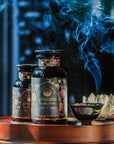Two dark glass jars labeled "Goddess of Earl: Lady Luck Oriental Beauty Oolong Tea" from Magic Hour are placed on a wooden surface. A small bowl releases smoke, contributing to the serene ambiance. The background is softly blurred, accentuating the aromatic setting of this organic loose leaf tea captured in the image.