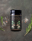 A black seasoning jar labeled "Lapsang Souchong Season Salt" sits on a dark textured surface, surrounded by rosemary sprigs, salt crystals, and peppercorns—perfect as a smoky mushroom seasoning or creative steak rub.