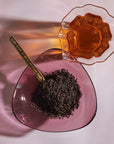 A spoon filled with loose Organic Lapsang Souchong: Smoky Black Tea rests on a pink, heart-shaped glass dish. A clear, patterned glass cup containing the smoked black tea is placed beside the dish, casting a shadow on the surface. The tea has a rich amber color and a distinct smoky flavor.