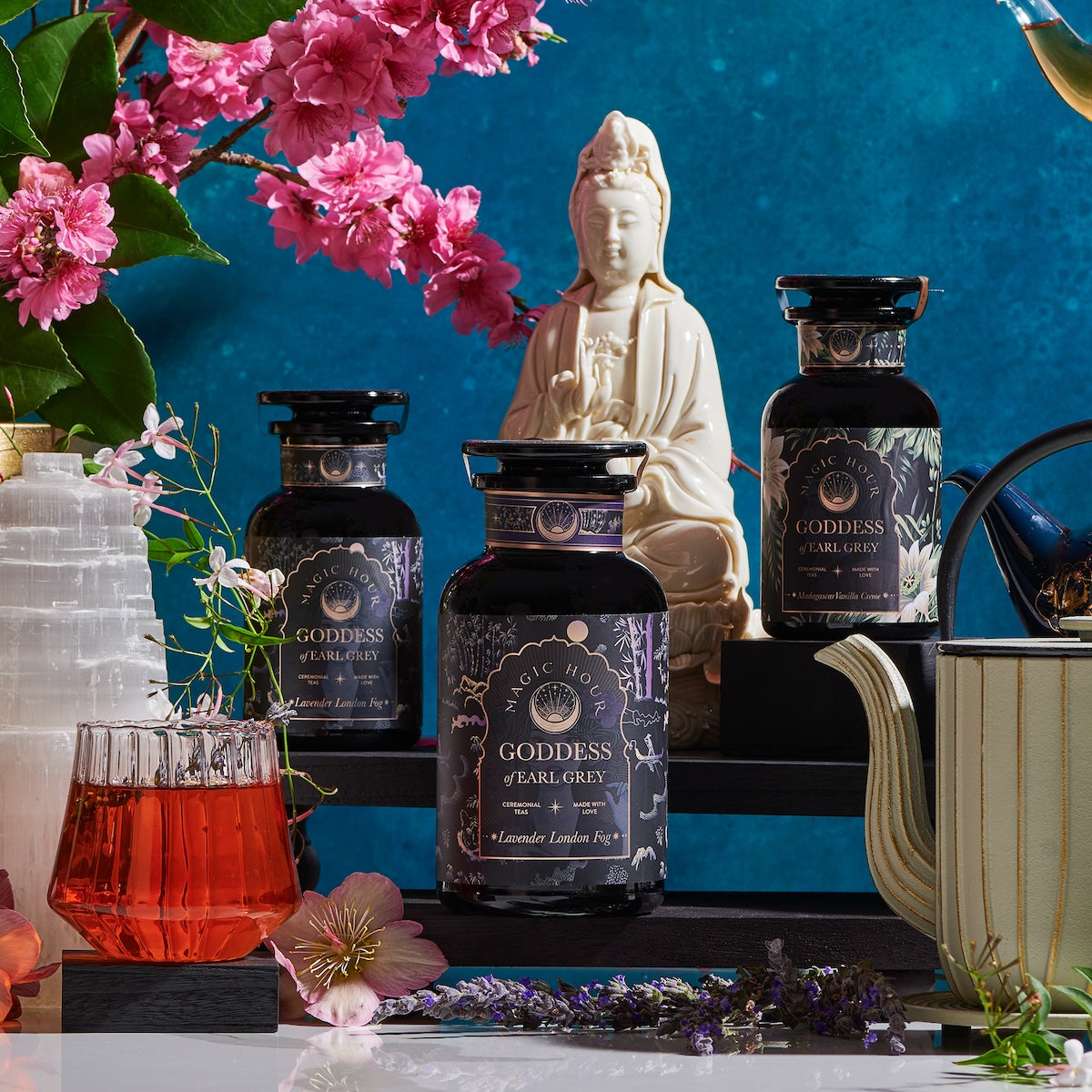 A serene display features three "Goddess of Earl: Lavender London Fog- Tea for Blooming Clarity & Calm Moods" jars by Magic Hour, a red glass of tea, and a teapot set against a blue backdrop. Pink flowers and a white statue of a seated figure complete the tranquil scene. A white crystal, decorative tea accessories, and the aromatherapeutic benefits of Lavender Earl Grey enhance relaxation and calm focus.