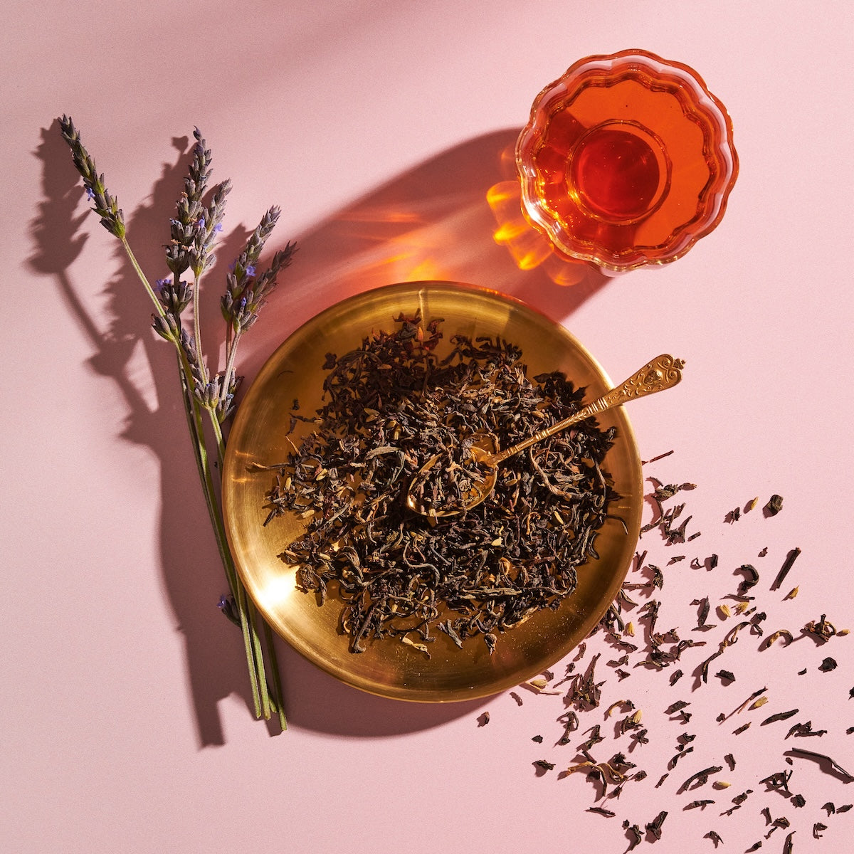 A brass plate filled with loose tea leaves and a brass spoon rests on a pink background. Next to the plate is a glass cup of Magic Hour&#39;s Goddess of Earl: Lavender London Fog - Tea for Blooming Clarity &amp; Calm Moods, with some lavender sprigs lying adjacent. Tea leaves are scattered around the plate, enhancing the scene&#39;s relaxation and calm focus.