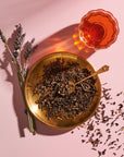 A brass plate filled with loose tea leaves and a brass spoon rests on a pink background. Next to the plate is a glass cup of Magic Hour's Goddess of Earl: Lavender London Fog - Tea for Blooming Clarity & Calm Moods, with some lavender sprigs lying adjacent. Tea leaves are scattered around the plate, enhancing the scene's relaxation and calm focus.