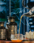 A stream of tea pours from a black teapot into a glass cup, beside two Goddess of Earl: Lavender London Fog tins and a white lotus candle on wood, all surrounded by rising steam for blooming clarity and calm moods.