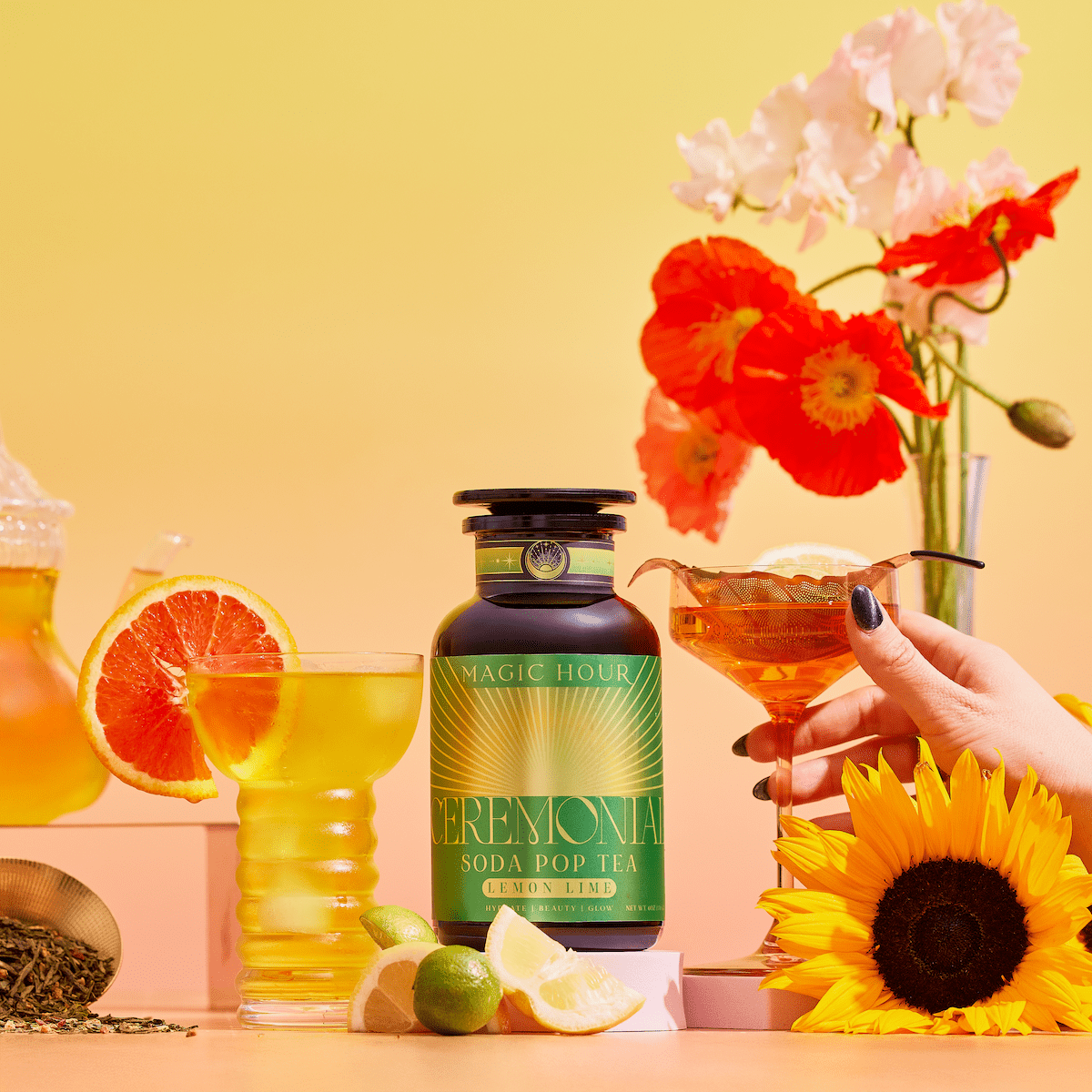 A bottle of Lemon Lime Green Tea: Ceremonial Soda Pop Tea with Sugar &quot;Destroyer&quot; sits among glasses of iced tea, green tea leaves, grapefruit, limes, a sunflower, lemon slices, and colorful flowers on a pastel yellow-orange background.