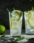 Two glasses of Magic Hour's Lemon Lime Green Tea, a ceremonial soda pop tea with Sugar "Destroyer" and organic ingredients, garnished with slices of lemon and mint leaves, sit on a textured surface with water droplets shimmering in the background. A whole lime and scattered mint leaves are in the foreground.