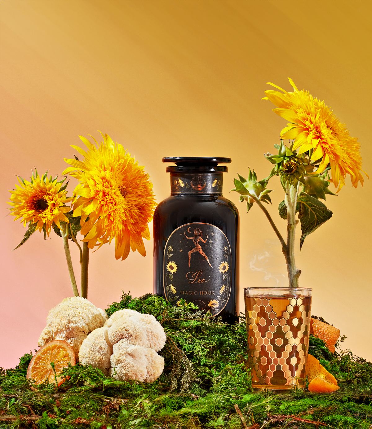 A still life composition featuring a black jar labeled "Leo Magic Hour" amidst vibrant yellow sunflowers and moss. A glass of tea with a decorative pattern is placed beside the jar, complemented by orange slices, white mushrooms, and green herbs.