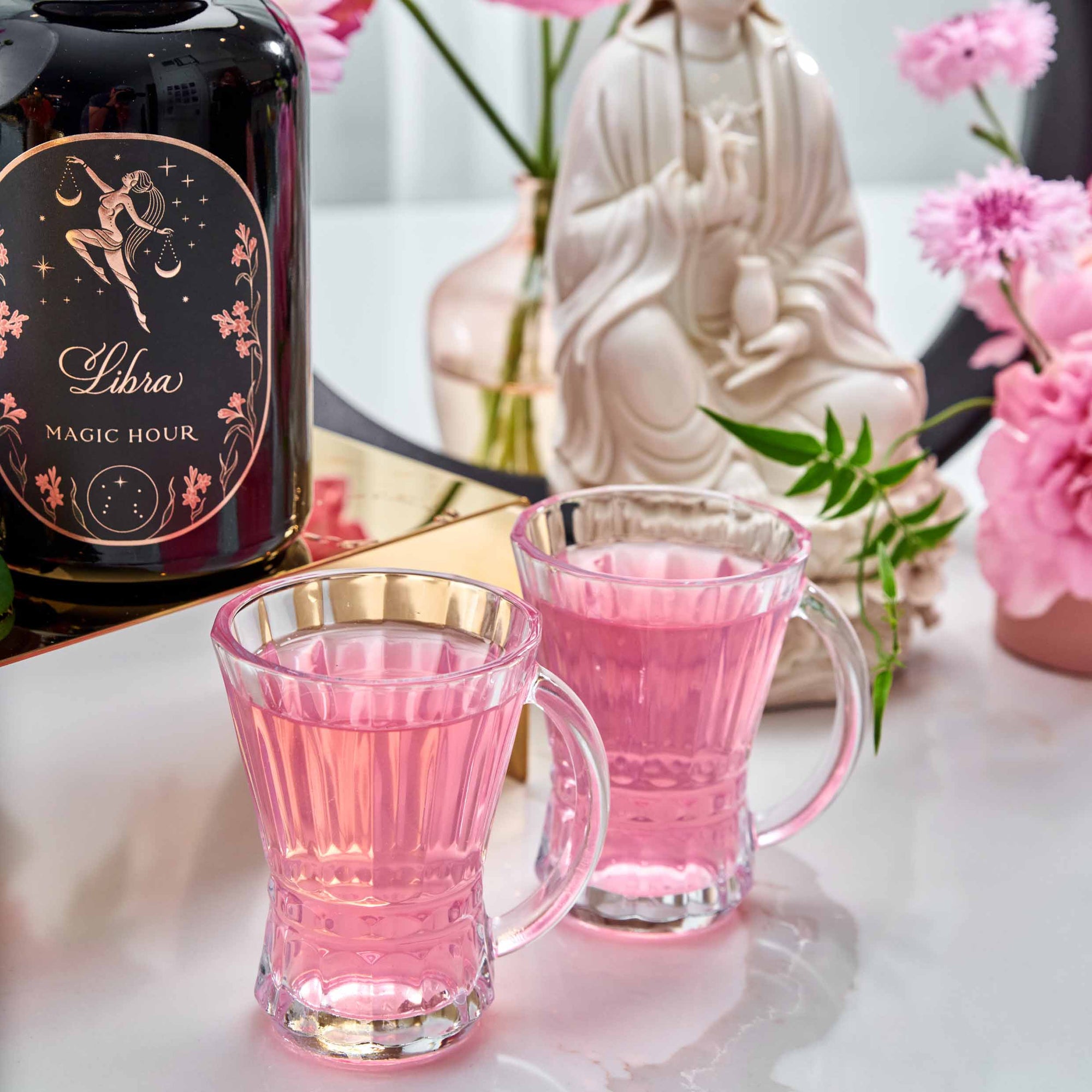Two clear glass mugs filled with Libra Shatavari-Cardamom-Rose White Love Cake Tea sit on a white surface by a black "Libra Magic Hour" tin, surrounded by pink flowers and a white statue in the background.
