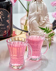 Two clear glass mugs filled with Libra Shatavari-Cardamom-Rose White Love Cake Tea sit on a white surface by a black "Libra Magic Hour" tin, surrounded by pink flowers and a white statue in the background.