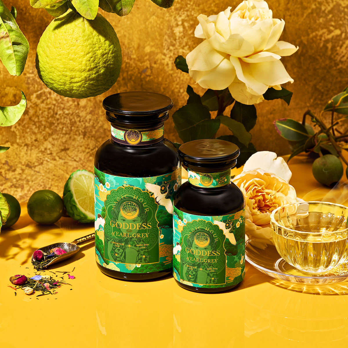 Two ornate green tea jars labeled "London Fog Green" sit amid fresh limes, roses, and tea leaves. A clear glass teacup filled with tea rests on a vibrant yellow surface, exuding floral complexity against a gold textured background.