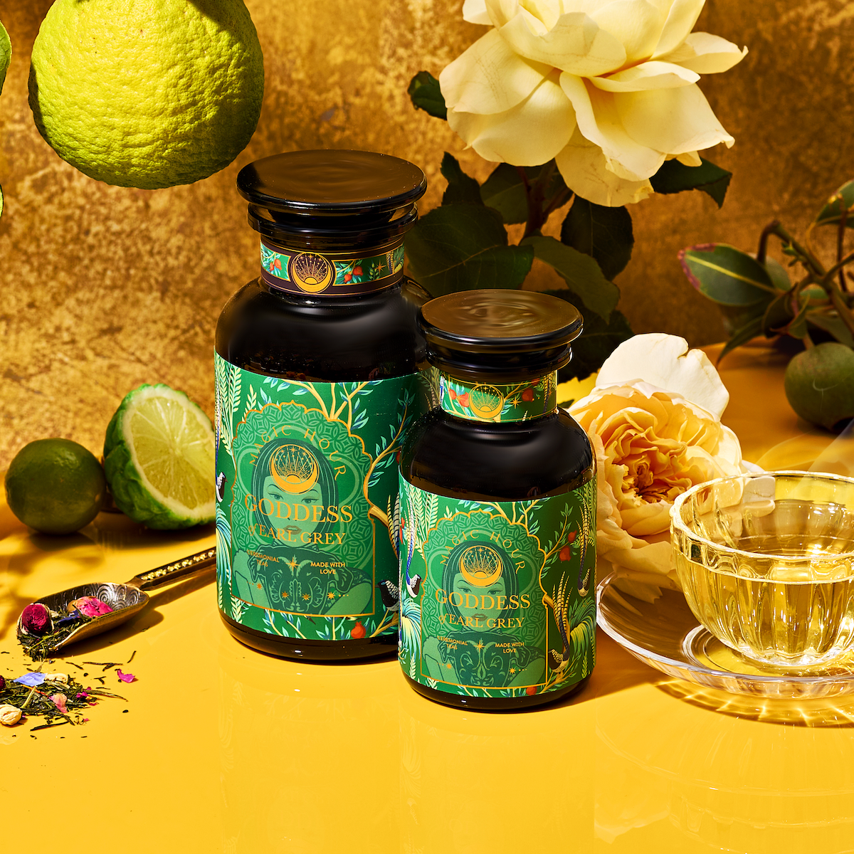 Two bottles of London Fog Oolong tea, with green and gold labels and hints of bergamot, sit amid a clear teacup, lime, roses, spoon, and loose tea. A bright yellow background enhances the vibrant natural theme.