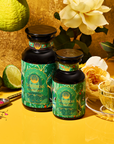 Two bottles of London Fog Oolong tea, with green and gold labels and hints of bergamot, sit amid a clear teacup, lime, roses, spoon, and loose tea. A bright yellow background enhances the vibrant natural theme.