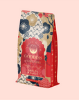 The "London Fog Rooibos" tea package features vibrant red, gold, and navy floral and celestial designs on a light pink background. Infused with aromatic bergamot essence, it offers a delightful blend of flavors.