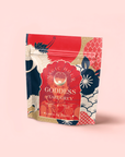 A colorful pouch of Magic Hour London Fog Rooibos tea with bold red, gold, blue, and cream floral patterns stands on a light pink surface—featuring hints of bergamot for a vibrant twist. The brand and tea name are displayed on the front.
