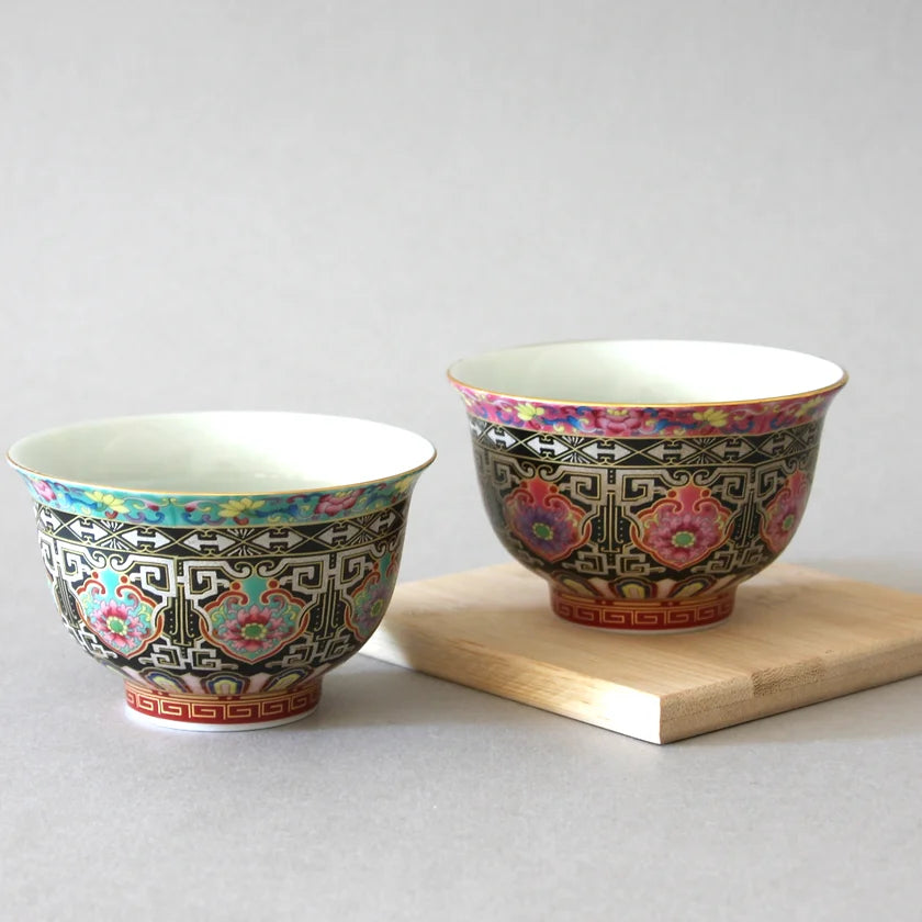 Two intricately decorated ceramic bowls from the Eastern Elm Longevity Cup Set of 2 sit on a wooden surface against a neutral background. One bowl features a black and blue design, while the other showcases a black and red design. Reflecting traditional Chinese porcelain, both have vivid, colorful accents.