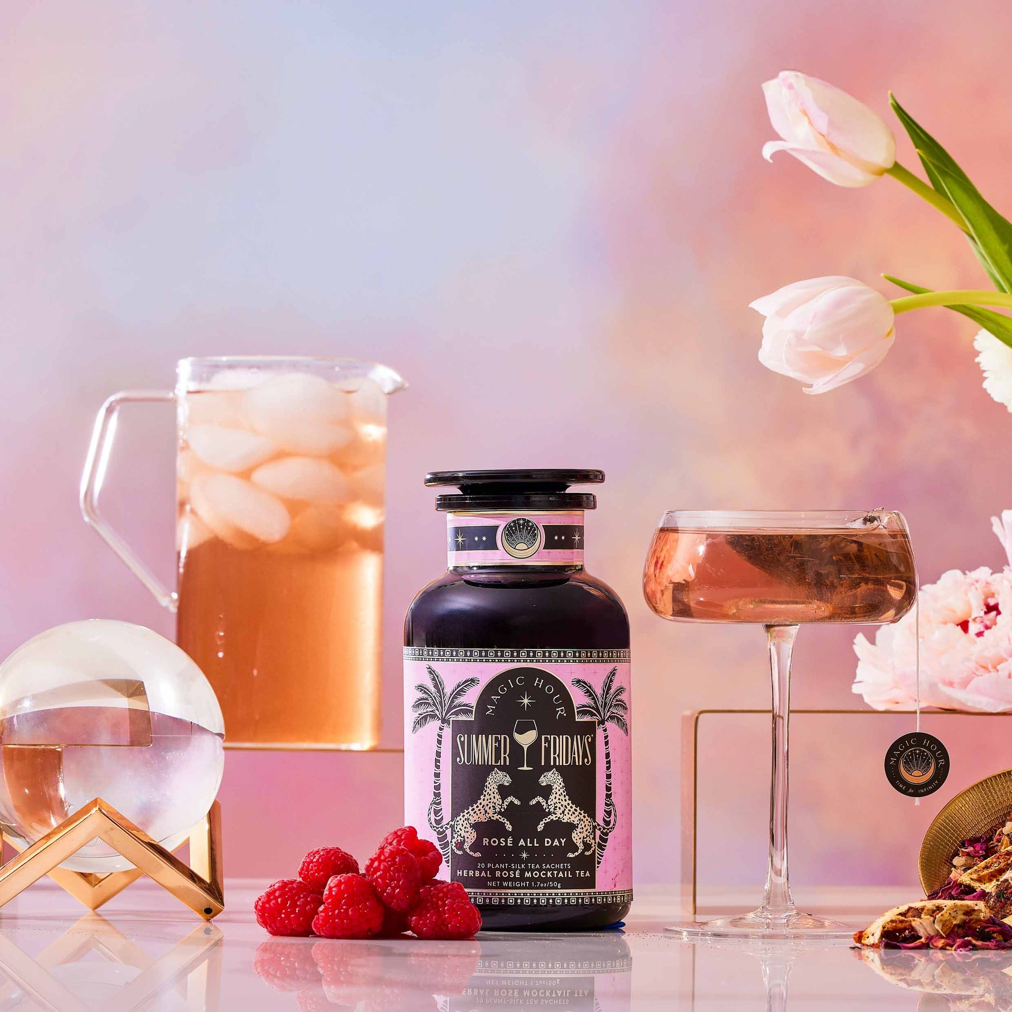 A bottle labeled "Summer Fridays™: Rose All Day," an alcohol-free rosé alternative, sits on a table with raspberries, a pink drink in a glass, iced beverage pitcher, crystal ball, and white tulips against a pastel background.