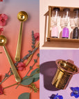 A composition featuring two gold measuring spoons, a trio of vibrant sand timers, and a gold tea infuser included in the Loose Leaf Tea Starter Set. The backdrop is adorned with pink, purple, and blue flowers on a gradient surface shifting from pink to purple. Ideal for enhancing the tea-making experience.