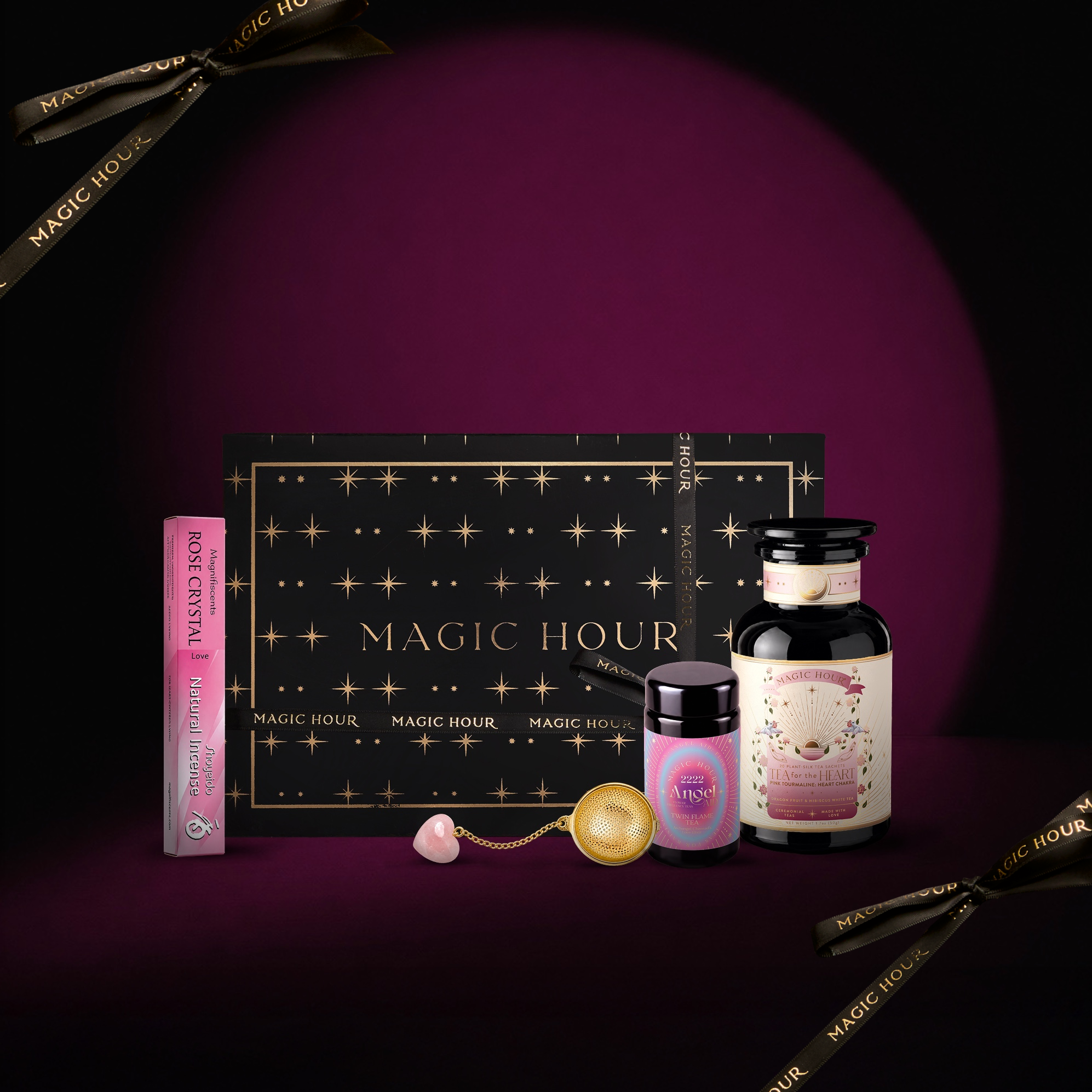 Introducing the Love Spell Box: a collection of luxury tea products and accessories featuring a black and gold “Magic Hour” box, an amethyst apothecary jar, a dropper bottle, a tea infuser, a heart-shaped crystal, and pink lip balm—perfect for love spells—all beautifully displayed on a purple background.