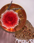 A red drink in a decorative glass sits on a round golden plate, garnished with two lavender sprigs. Beside it rest dried botanicals from Tea Discovery Club's curated blends and a gold leaf accent on a light surface.