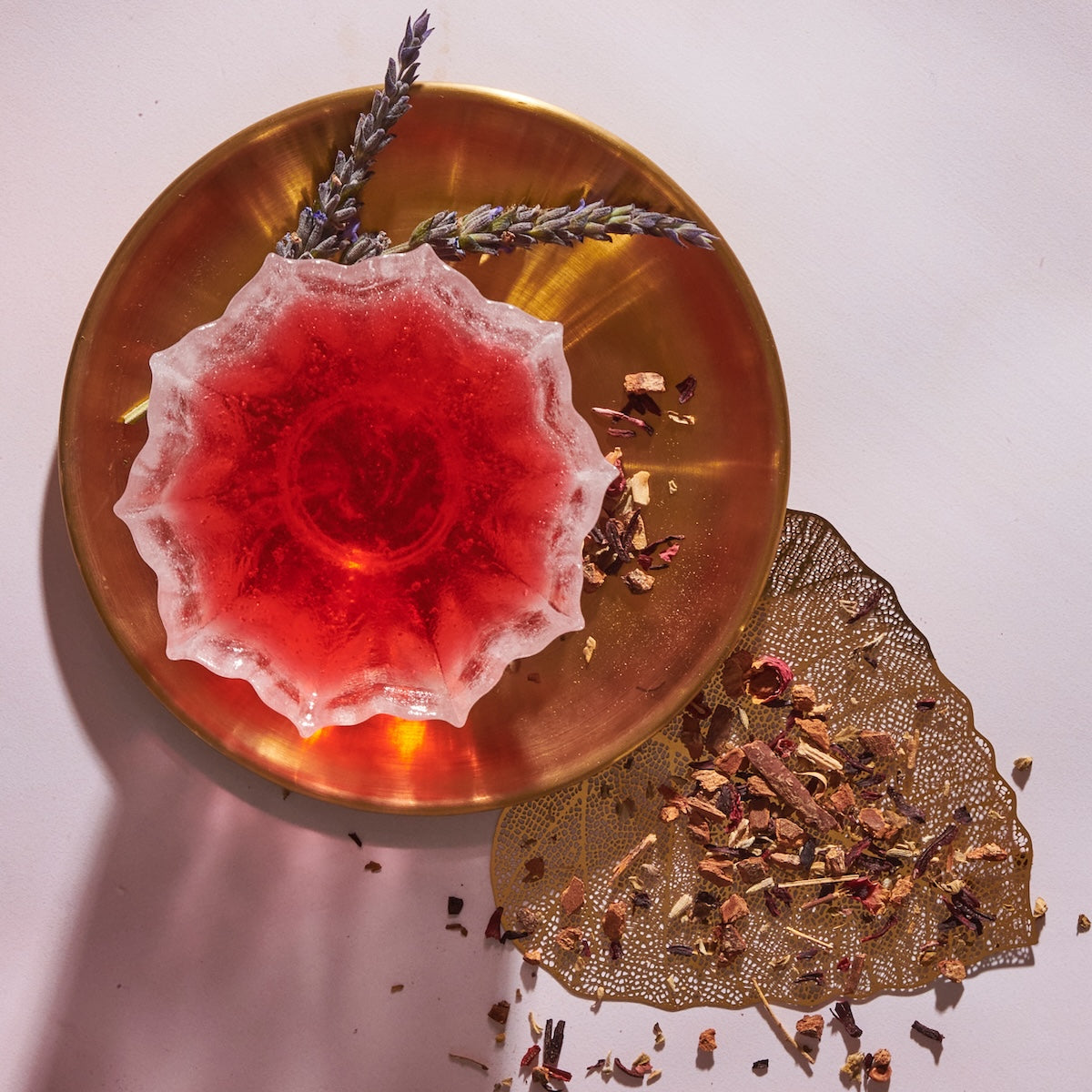 A red drink in a clear, decorative glass sits on a golden plate. The drink, infused with Magic Hour&#39;s The Lovers, is garnished with sprigs of lavender. Next to the plate are scattered pieces of dried herbs and a gold, lace-like leaf. The background is a light, neutral color.