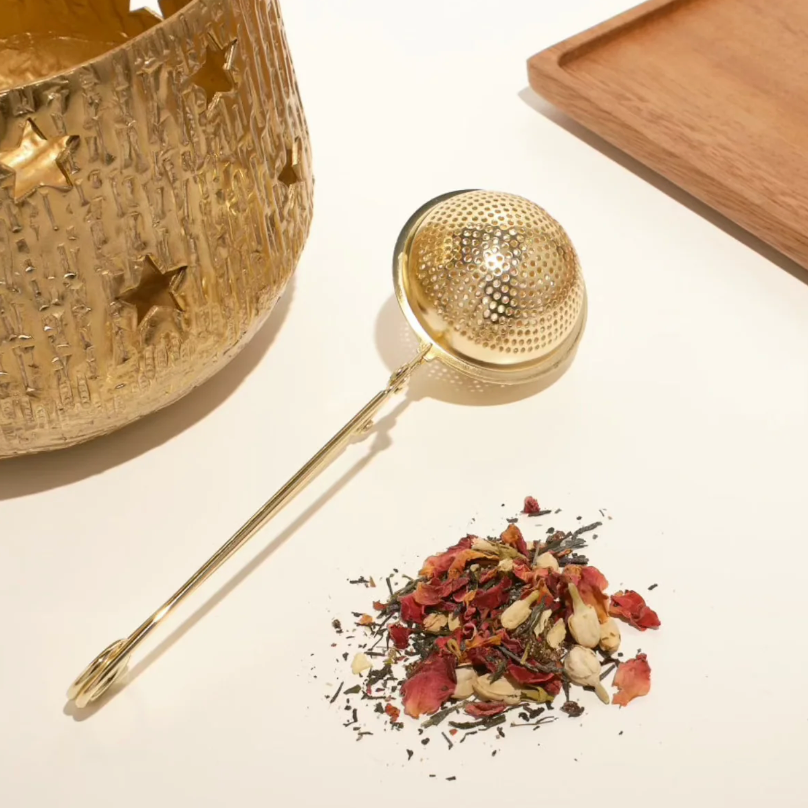 A golden tea infuser lies next to a small pile of Organic Tea leaves and flowers. An intricately designed golden container with star cutouts stands on the left, and a wooden tray is positioned in the background to the right, adding a touch of elegance to your Bestsellers of Magic Tea Starter Set- 60-75 cup sampler box by Magic Hour ritual.