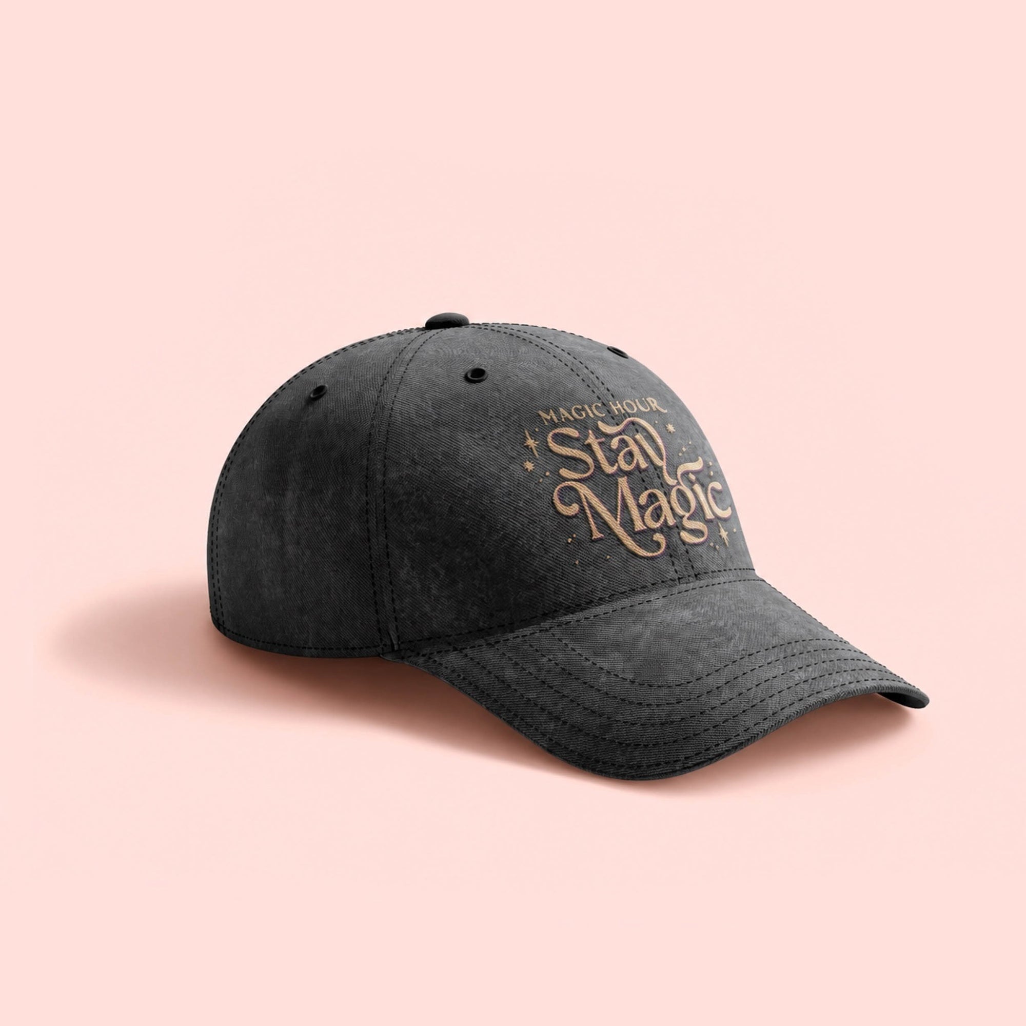 The Stay Magic Embroidered Dad Hat in dark gray washed cotton twill features “Magic Hour, Stay Magic” in decorative beige embroidery on the front and is shown against a light pink background.