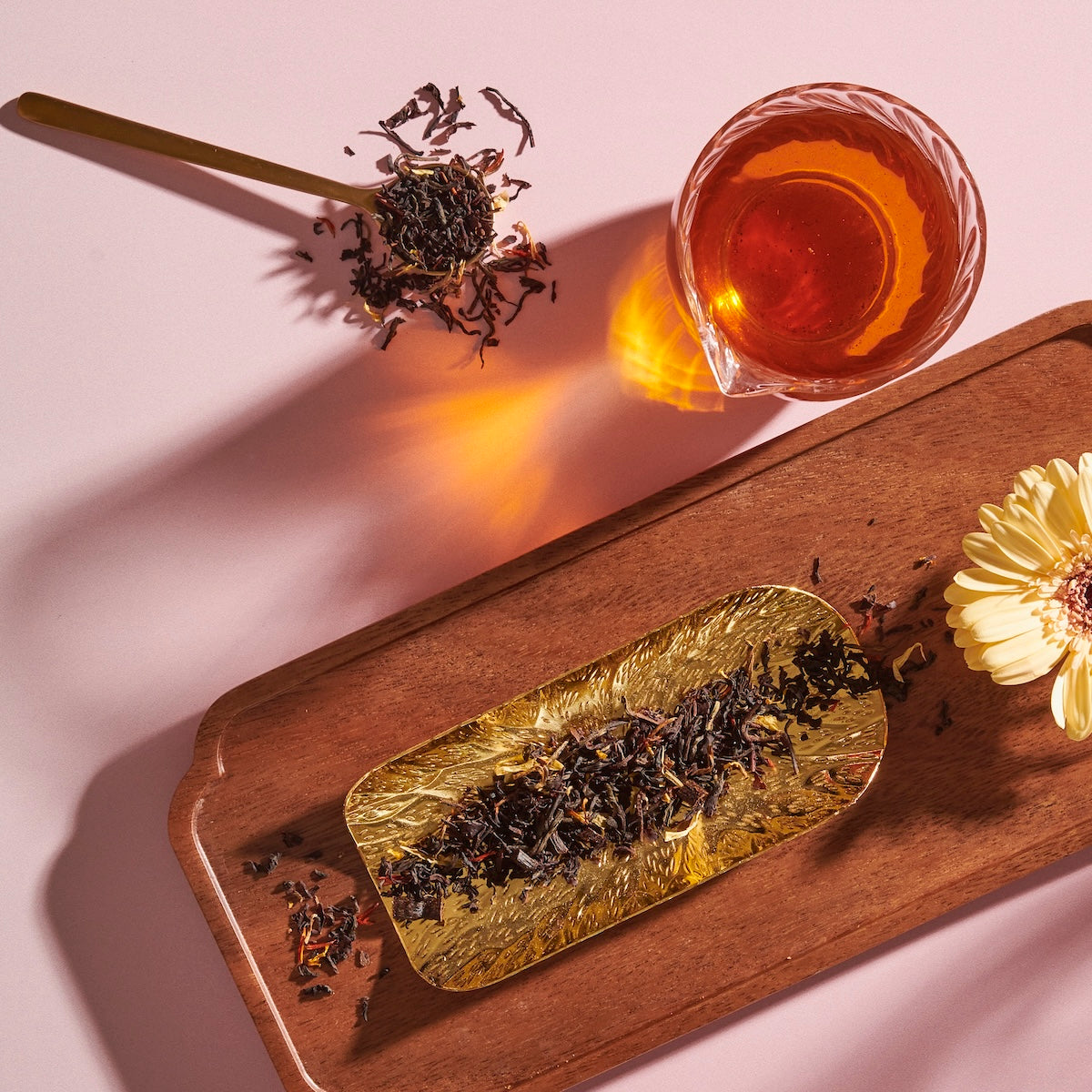 A flat lay image featuring a glass of Magic Hour&#39;s Goddess of Earl: Madagascar Vanilla Creme Tea for Soothing Delight &amp; Delicious Decadence, a spoon with loose tea leaves, and a golden tray with more tea leaves resting on a wooden tray. A single yellow flower decorates the corner of the wooden tray, all set against a light pink background.