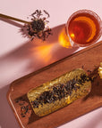 A flat lay image featuring a glass of Magic Hour's Goddess of Earl: Madagascar Vanilla Creme Tea for Soothing Delight & Delicious Decadence, a spoon with loose tea leaves, and a golden tray with more tea leaves resting on a wooden tray. A single yellow flower decorates the corner of the wooden tray, all set against a light pink background.