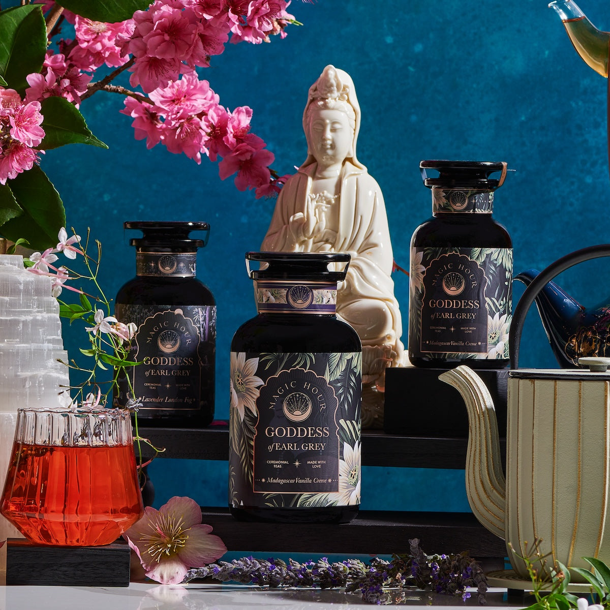 A serene tea setup featuring three &quot;Goddess of Earl: Madagascar Vanilla Creme Tea for Soothing Delight &amp; Delicious Decadence&quot; tea jars from Magic Hour, a white statue, blooming pink flowers, a white striped teapot, and a glass of black tea on a wooden table. A calming blue backdrop enhances the tranquil scene.