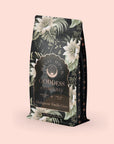 A decorative tea package features a soft pink floral design with the label "Madagascar Vanilla Earl Grey Creme Tea," highlighting notes of Madagascar Vanilla Creme and organic black tea, complemented by intricate patterns and vanilla orchid hints.