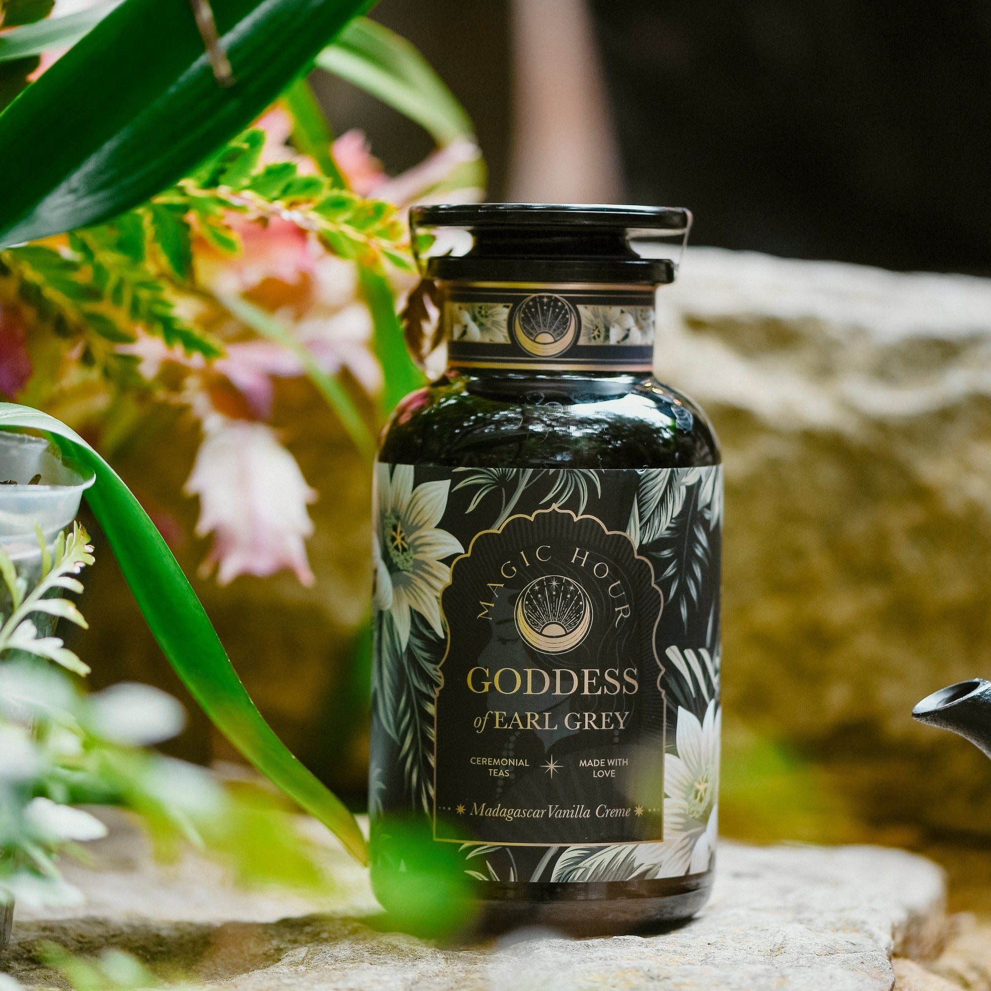 A black glass jar labeled "Goddess of Earl: Madagascar Vanilla Creme Tea for Soothing Delight &amp; Delicious Decadence" rests on stone, surrounded by green leaves and pink flowers—a serene scene for lovers of organic black tea.