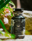 A black glass jar labeled "Goddess of Earl: Madagascar Vanilla Creme Tea for Soothing Delight & Delicious Decadence" rests on stone, surrounded by green leaves and pink flowers—a serene scene for lovers of organic black tea.