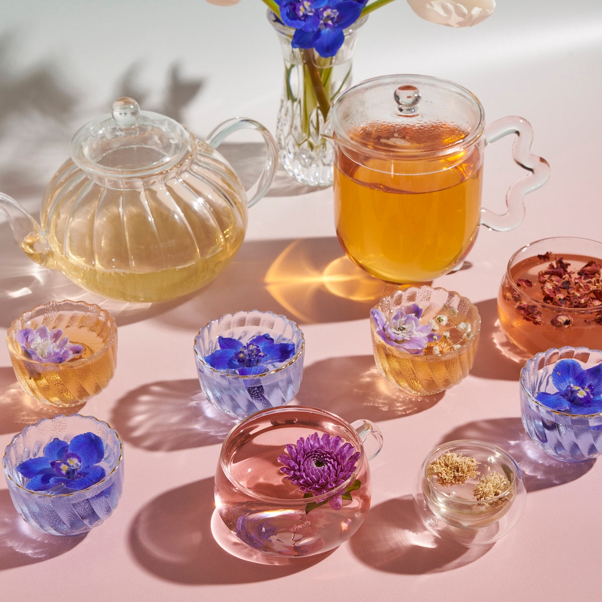 A display featuring clear glass teacups and teapots beautifully highlights the vibrant colors of The Best of Spring Box teas, with some cups adorned with floating flowers. The arrangement includes purple and blue flowers inside the teacups, complemented by a bouquet in the background, all set against a light pink surface.