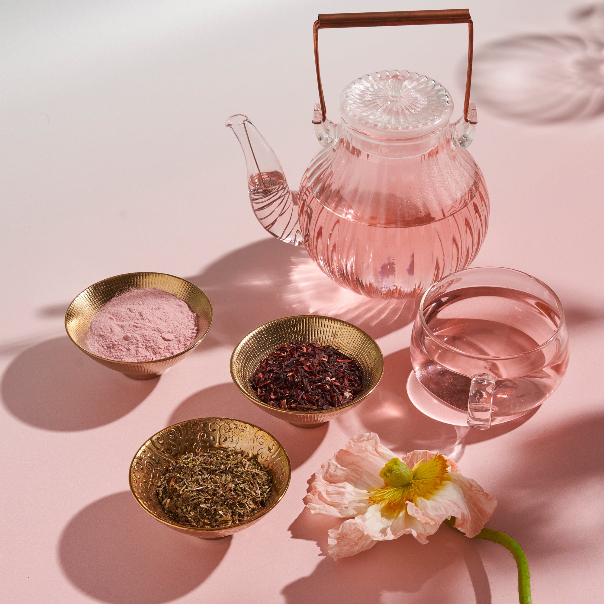 A glass teapot with pink liquid, a glass teacup, and three brass bowls holding pink powder, dried herbs, and tea leaves from The Best of Spring Box are displayed on a light pink surface with a pale flower nearby.