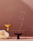 A minimalist scene featuring Magic Hour's high-quality Ceremonial Incense Burners in brass and black, with lit Japanese incense sticks emitting smoke. The brass holder is elevated on a metal stand. A single white rose lies beside them against a neutral tan background.