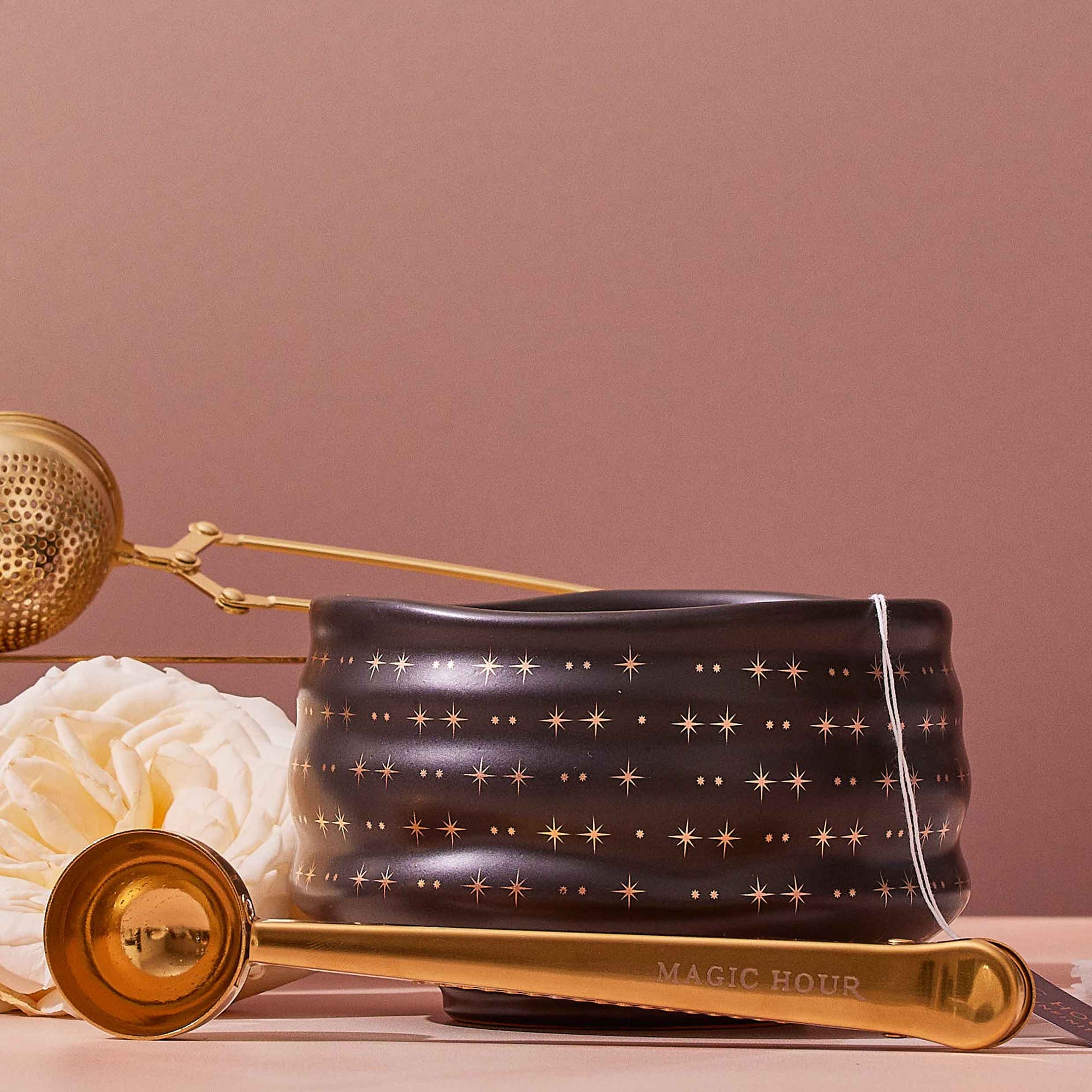 A Ceremonial Matcha Bowl features 14k gold star detailing, accompanied by a gold “Magic Hour” teaspoon, a tea infuser, a white rose, and a teabag, all arranged on a soft pink background.