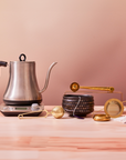 The Magic Hour Starter Kit by Magic Hour rests elegantly on a wooden surface, ideal for an exquisite tea ceremony. The kit includes a silver gooseneck electric kettle with a black handle, perfectly complemented by a black dotted teapot featuring an orange interior, artisan-crafted gold strainers, a tea scoop, and a small mirror. A beige wall provides the backdrop to this serene setting.