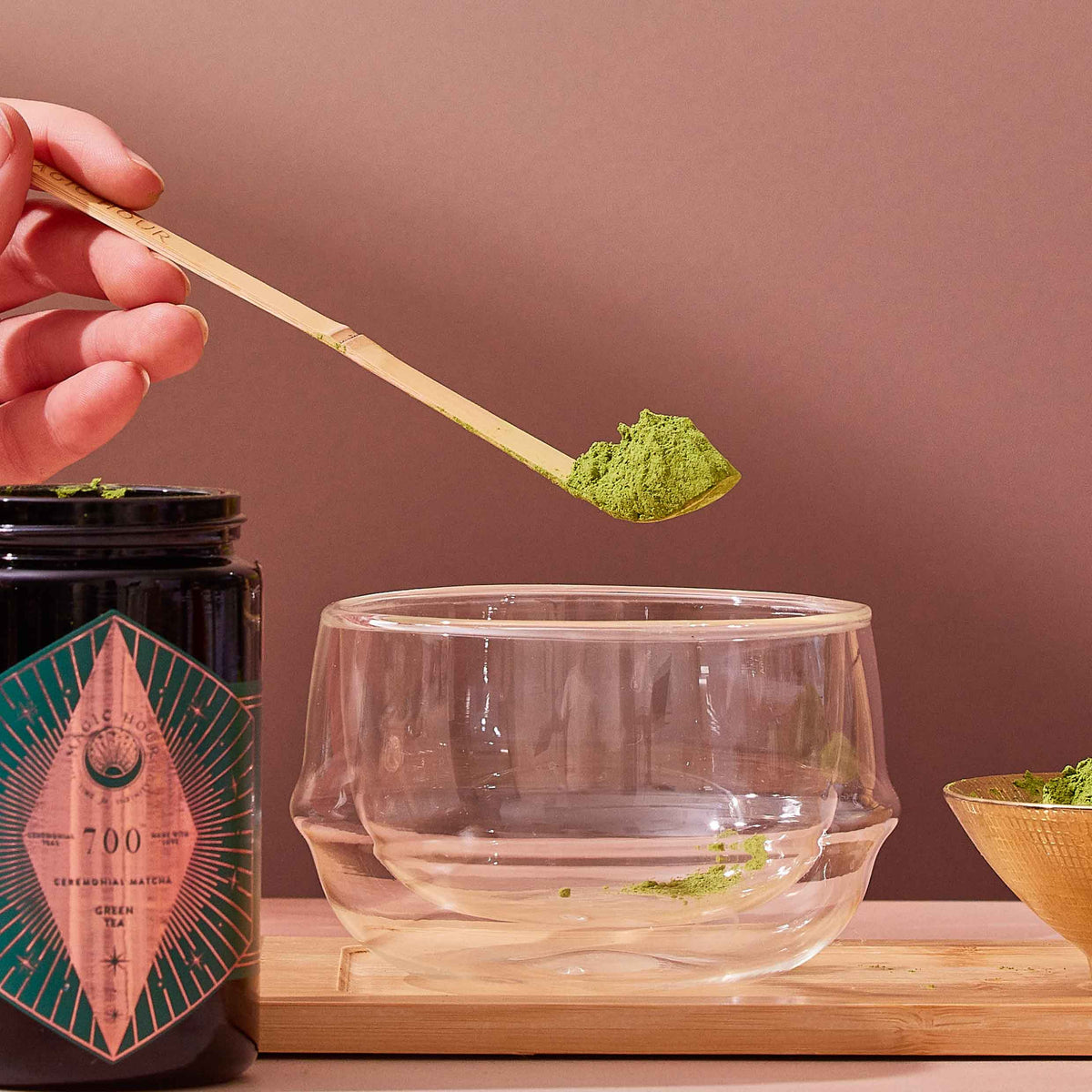 A hand holds the Ceremonial Matcha Bamboo Scoop filled with vibrant green matcha powder above a clear double-walled glass cup. A matching matcha jar and bowl, each holding more powder, rest on a wooden tray, capturing the serene atmosphere of a modern tea ceremony against a neutral backdrop.