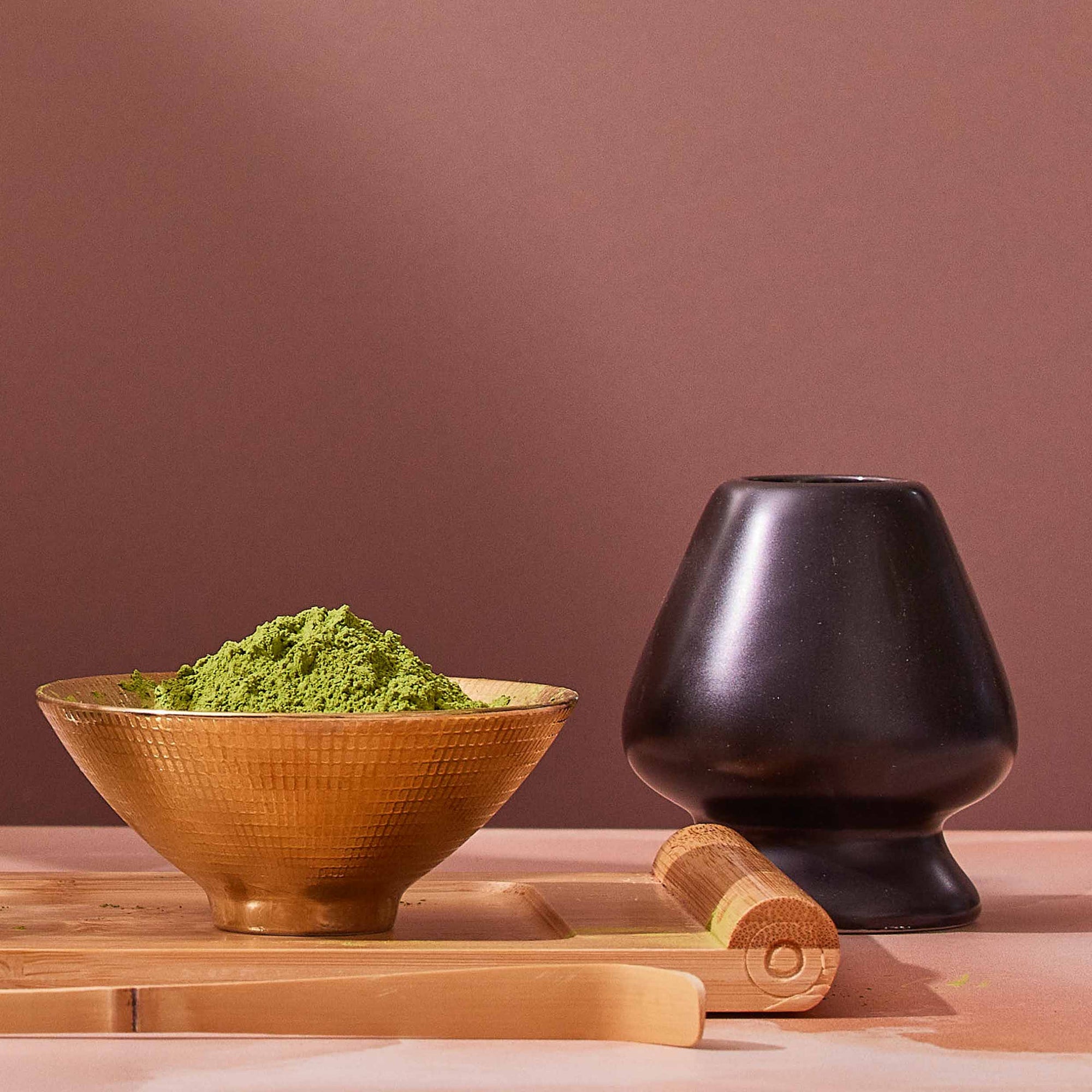 A gold bowl filled with green matcha powder sits next to a Matte Black Chasen - Matcha Whisk Stand, a bamboo whisk, and a bamboo scoop on a wooden tray against a mauve background, evoking the tranquility of a Japanese tea ceremony.