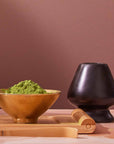 A gold bowl filled with green matcha powder sits next to a Matte Black Chasen - Matcha Whisk Stand, a bamboo whisk, and a bamboo scoop on a wooden tray against a mauve background, evoking the tranquility of a Japanese tea ceremony.
