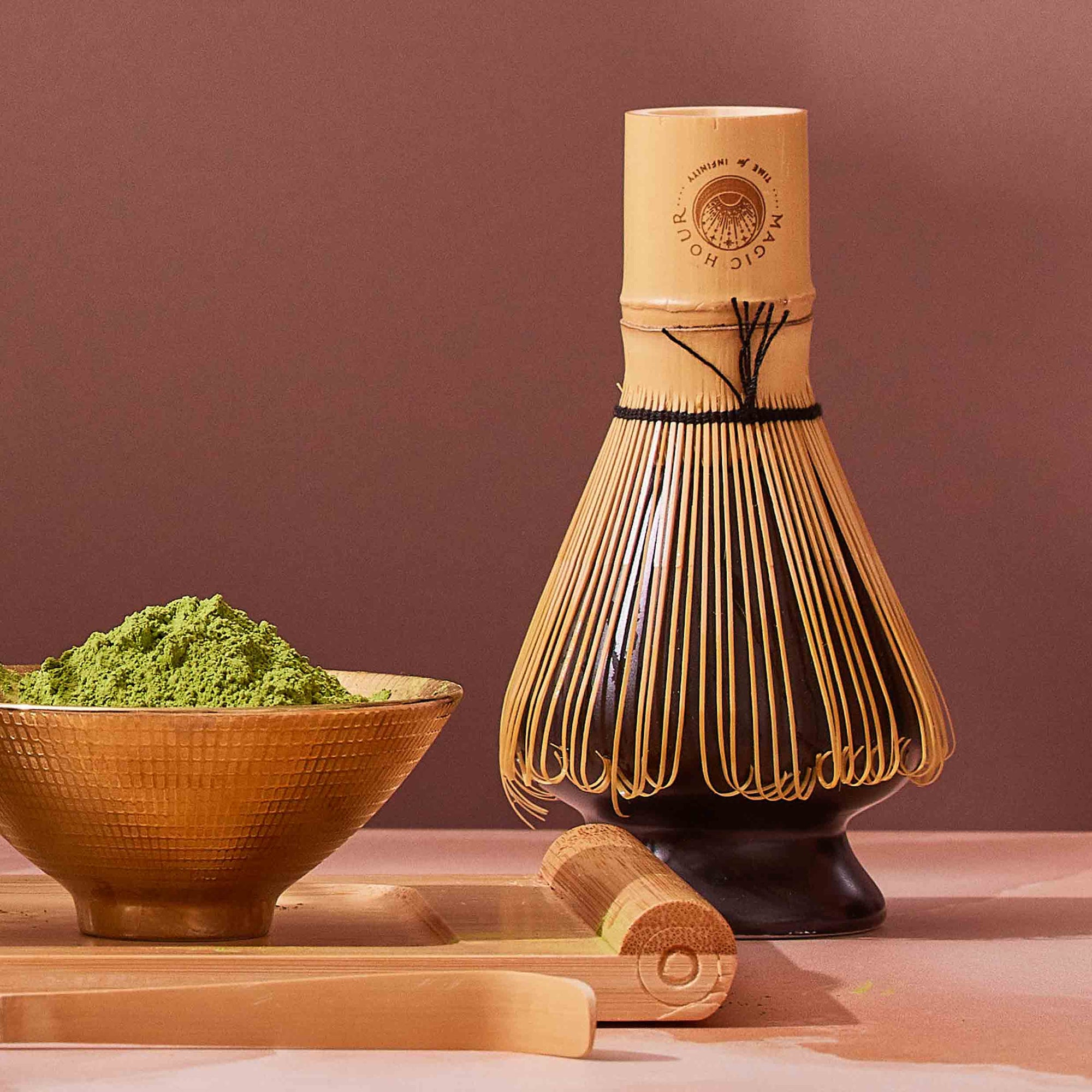 The Artisanal Magic Hour Matcha Whisk stands upright beside a bowl filled with vibrant green matcha powder and a bamboo scoop, all arranged on a light surface against a muted brown background, emphasizing the sustainable appeal of these handmade tea tools.
