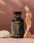 The World, a decorative black container adorned with floral motifs and illustrations, rests on a surface beside a small white chrysanthemum. Nearby, an intricately carved ivory-colored statuette of a woman enhances the elegance. The background displays a warm, muted brown hue.