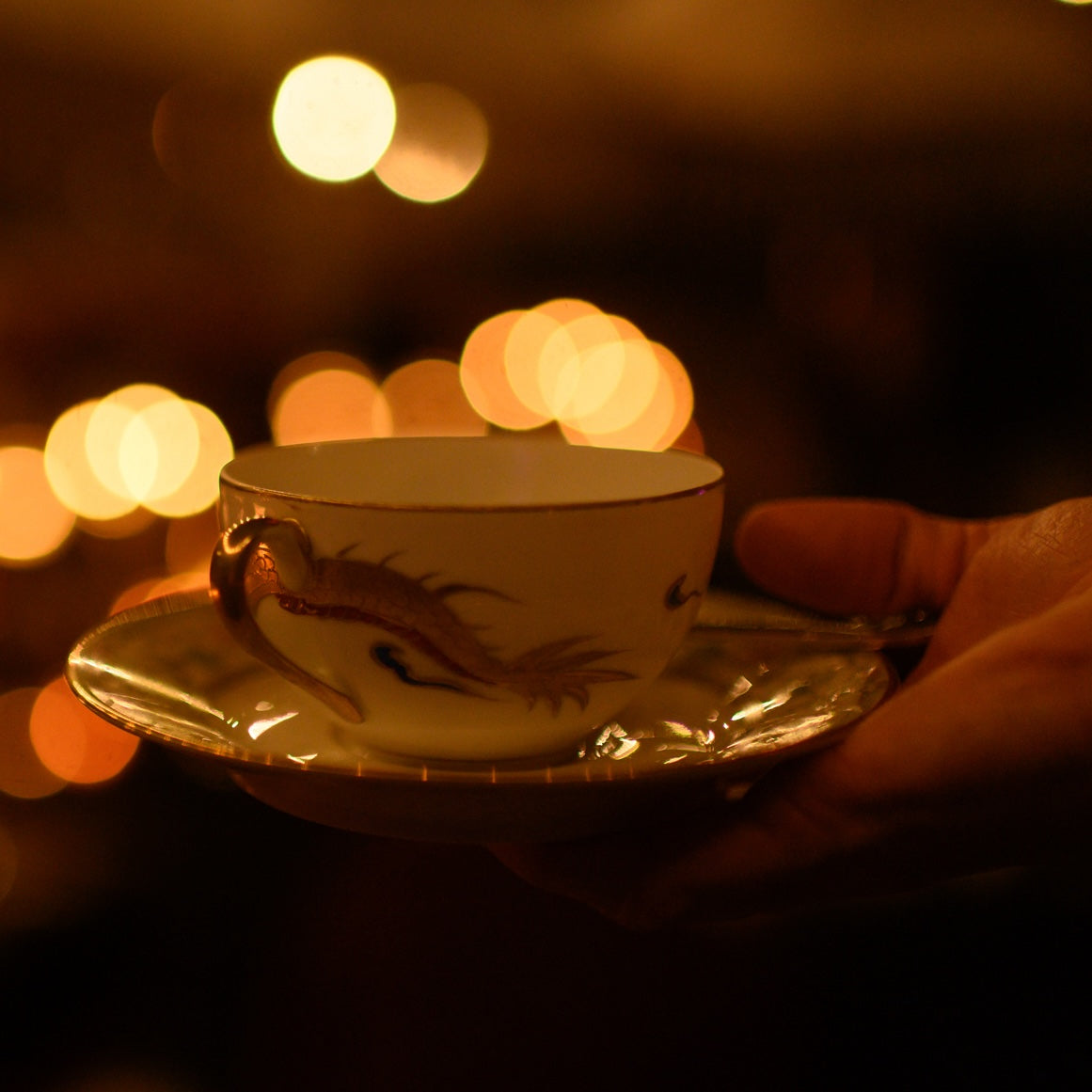 A hand holds a delicate porcelain teacup and saucer with a dragon design, set against a warm, softly lit background with blurred circular bokeh lights, capturing the essence of the Guiding Lightworker Circle: $500 Pledge at Club Magic Hour.