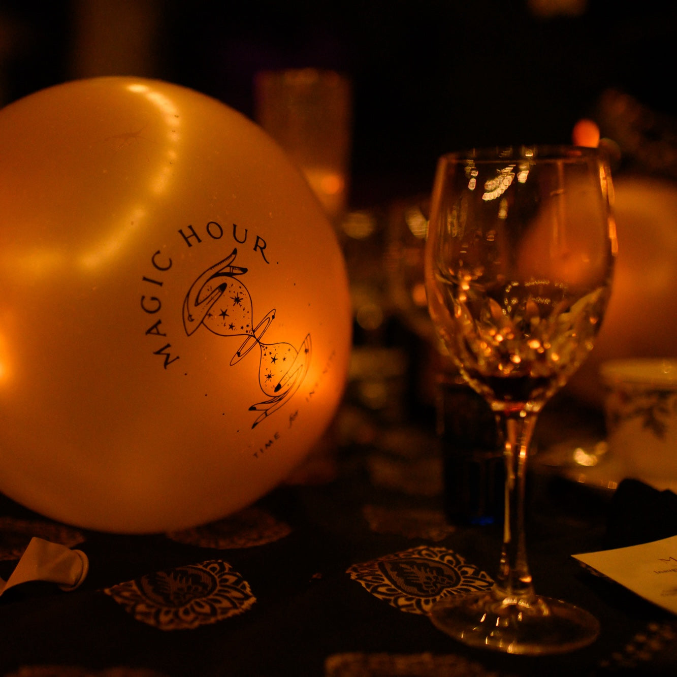 A dimly lit scene features a glowing balloon adorned with the &quot;Friend for Infinitea&quot; logo and an hourglass design. In the foreground, a partially filled wine glass sits elegantly on the table. The intricately patterned tablecloth adds a touch of sophistication to this exclusive $250 Pledge gathering by Club Magic Hour.