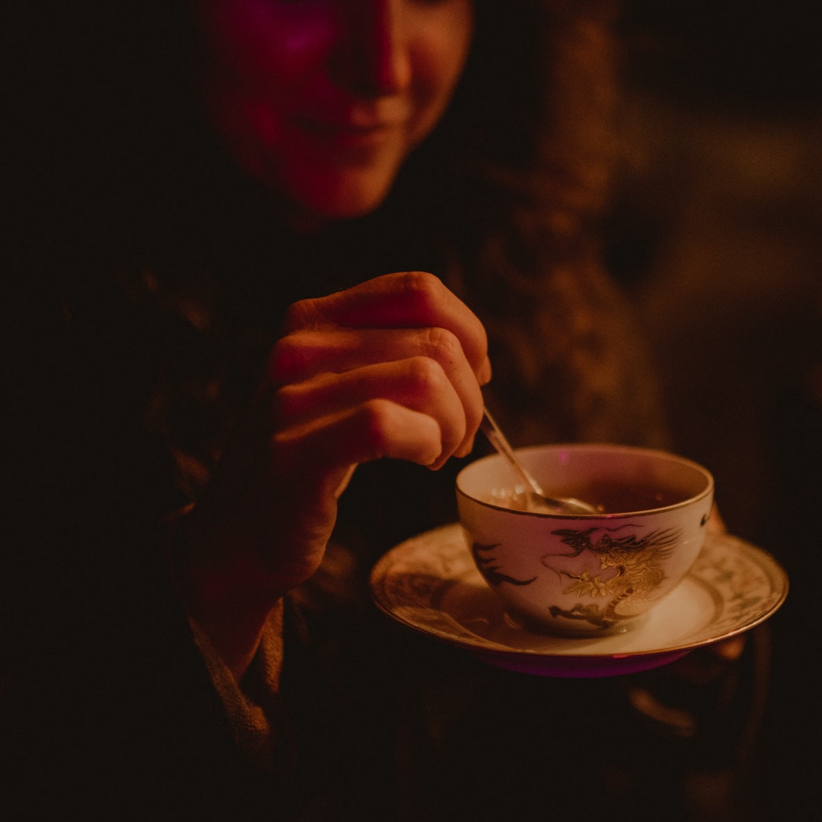 A person with long hair holds a decorated porcelain teacup in one hand and gently stirs the tea with a spoon in the other. The scene at Club Magic Hour is dimly lit, creating a cozy and intimate atmosphere perfect for this private event featuring the exclusive Founding Spirit Guide: $7,000 Pledge.